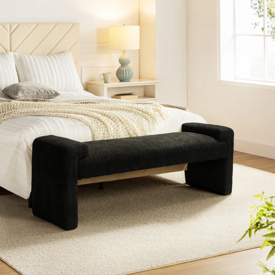 A bedroom scene with a beige bed featuring white and cream bedding, a black upholstered bench with dimensions 17.3'' H x 63'' W x 17.7'' D at the foot, a side table with a lamp and book, and a light-colored rug on a wooden floor.