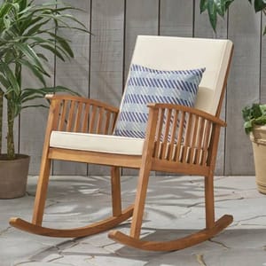 Wooden rocking chair with slatted armrests, a light beige cushion, and a decorative blue and white striped pillow. Surrounded by potted plants on a stone patio.