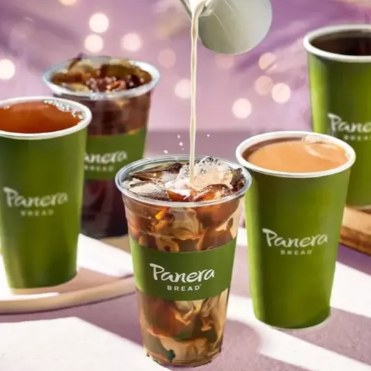 Panera Bread cups filled with various drinks, including iced beverages and coffee, feature prominently. Milk is being poured into a clear cup with ice and coffee. All cups have the Panera Bread logo.