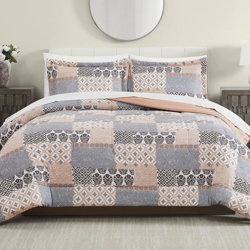 A bed is adorned with a patchwork-patterned quilt and matching pillow shams featuring shades of gray, peach, and white. The bedroom setting includes a circular mirror, side tables, and lamps.