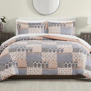 A bed is adorned with a patchwork-patterned quilt and matching pillow shams featuring shades of gray, peach, and white. The bedroom setting includes a circular mirror, side tables, and lamps.