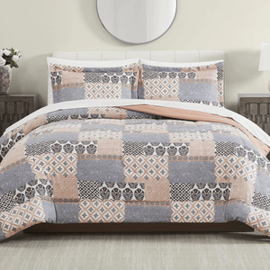 A bed is adorned with a patchwork-patterned quilt and matching pillow shams featuring shades of gray, peach, and white. The bedroom setting includes a circular mirror, side tables, and lamps.