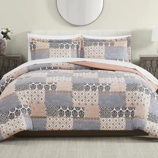 A bed is adorned with a patchwork-patterned quilt and matching pillow shams featuring shades of gray, peach, and white. The bedroom setting includes a circular mirror, side tables, and lamps.