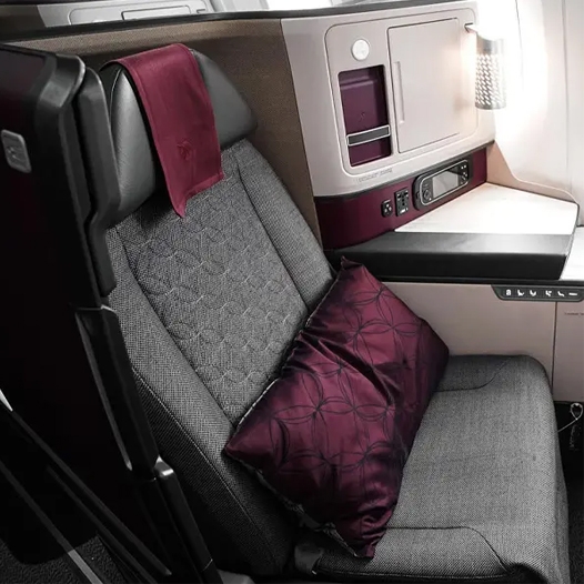 A luxurious airline seat featuring a textured grey fabric, a maroon pillow, and backrest cloth. The seat includes a built-in storage compartment, a personal light, and control buttons for adjustments, offering privacy and comfort.