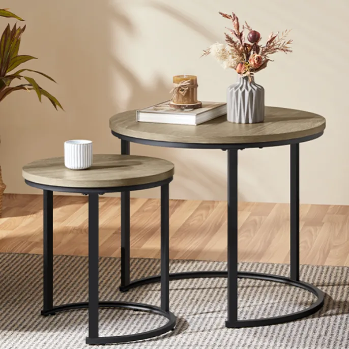 A set of two nesting round coffee tables with black metal legs and wooden tops, displayed on a striped rug. The larger table holds a vase with dried flowers and books with a candle, while the smaller one has a white mug.