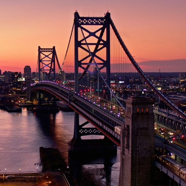 A suspension bridge spans a river at sunset, with vehicle and train traffic crossing between cityscapes illuminated by colorful lights.