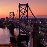 A suspension bridge spans a river at sunset, with vehicle and train traffic crossing between cityscapes illuminated by colorful lights.