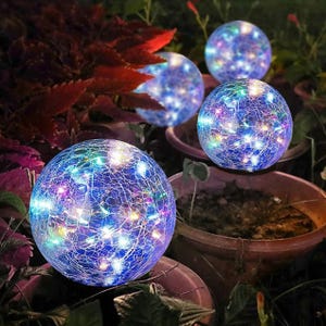 Four crackled glass solar LED globes with colorful lights are placed among garden plants.