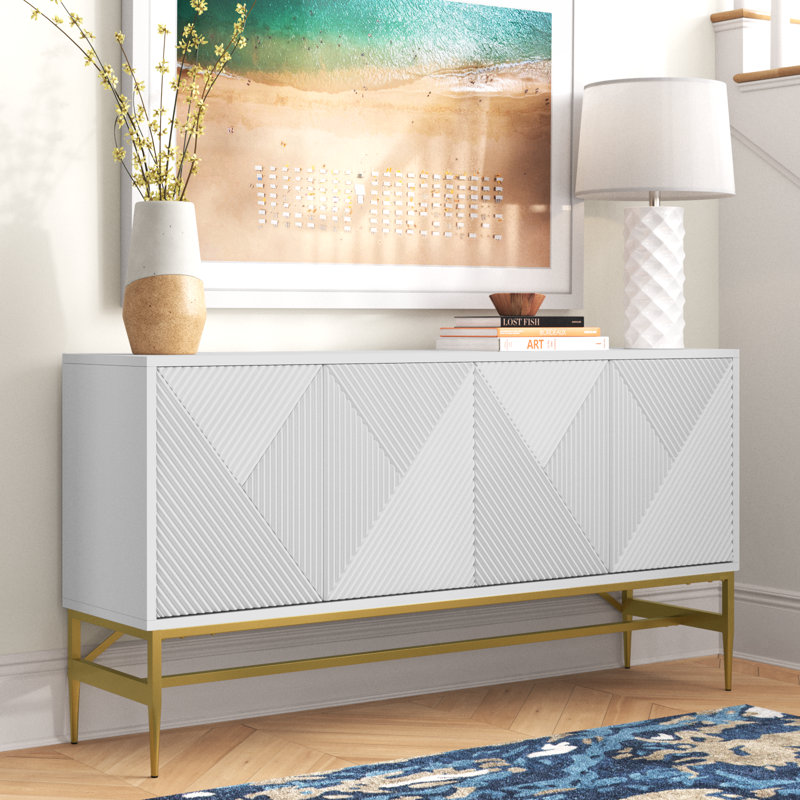 A white sideboard with a geometric pattern on its doors, featuring a brass-finished metal base, flanked by a vase, books, and a table lamp, against an artwork backdrop.