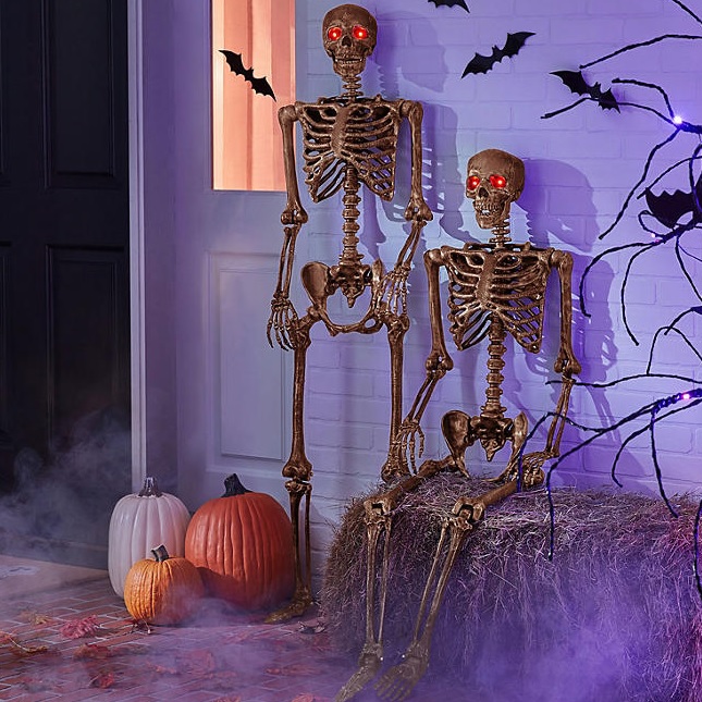 Two bronze-colored skeletal figures with red illuminated eyes are posed standing; one is smaller, suggesting a child, set amidst Halloween-themed decor.