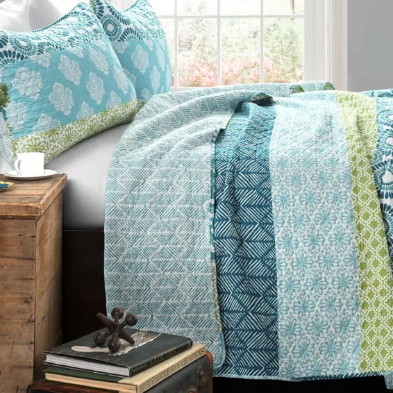 A bed with a colorful quilt featuring blue and green patterns, alongside matching pillowcases. A rustic wooden nightstand holds a book and an ornament, and a window provides natural light.