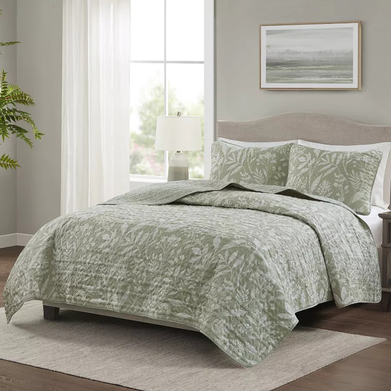 A bed is adorned with a green quilt featuring a leafy pattern, accompanied by matching pillow shams. A lamp and framed artwork complement the neutral-toned bedroom decor.