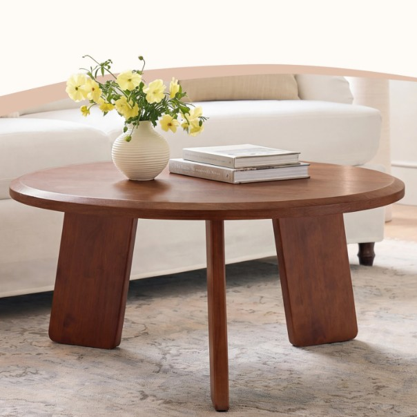 A round wooden coffee table with a unique three-legged design, featuring a vase with yellow flowers and two stacked books on top, situated in a living room setting.