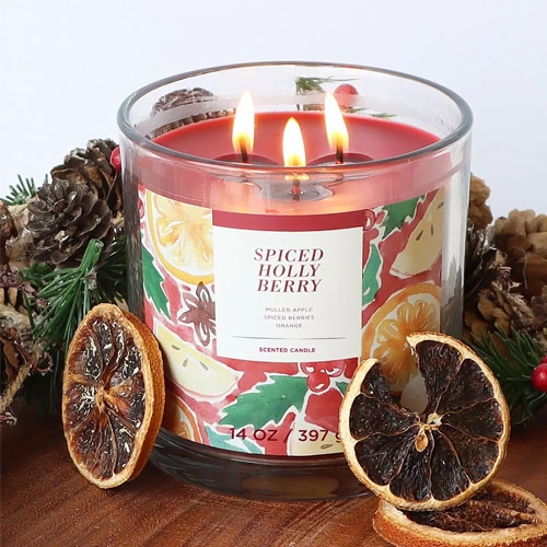 a red three wick candle surrounded by pine cones and dried oranges