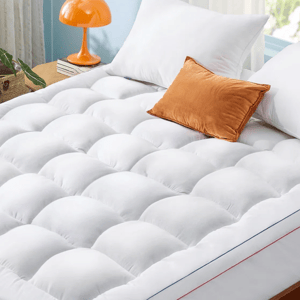 Plush mattress topper on a bed with two white pillows and one orange pillow. An orange lamp and books are on the bedside table.
