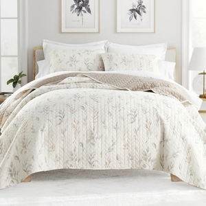 A neatly made bed features a quilt with a subtle floral pattern, complemented by matching pillows. The room includes light gray and beige tones, with wall art and a lamp enhancing the calm ambiance.