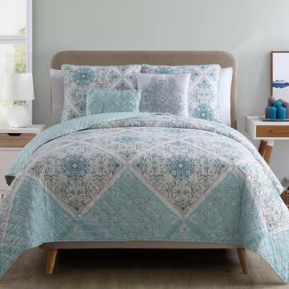A bed features a light blue and white patterned quilt with matching pillow shams. The setup includes a bedside table with a lamp on the left and candles and artwork on the right.