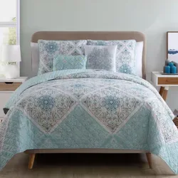 A bed features a light blue and white patterned quilt with matching pillow shams. The setup includes a bedside table with a lamp on the left and candles and artwork on the right.