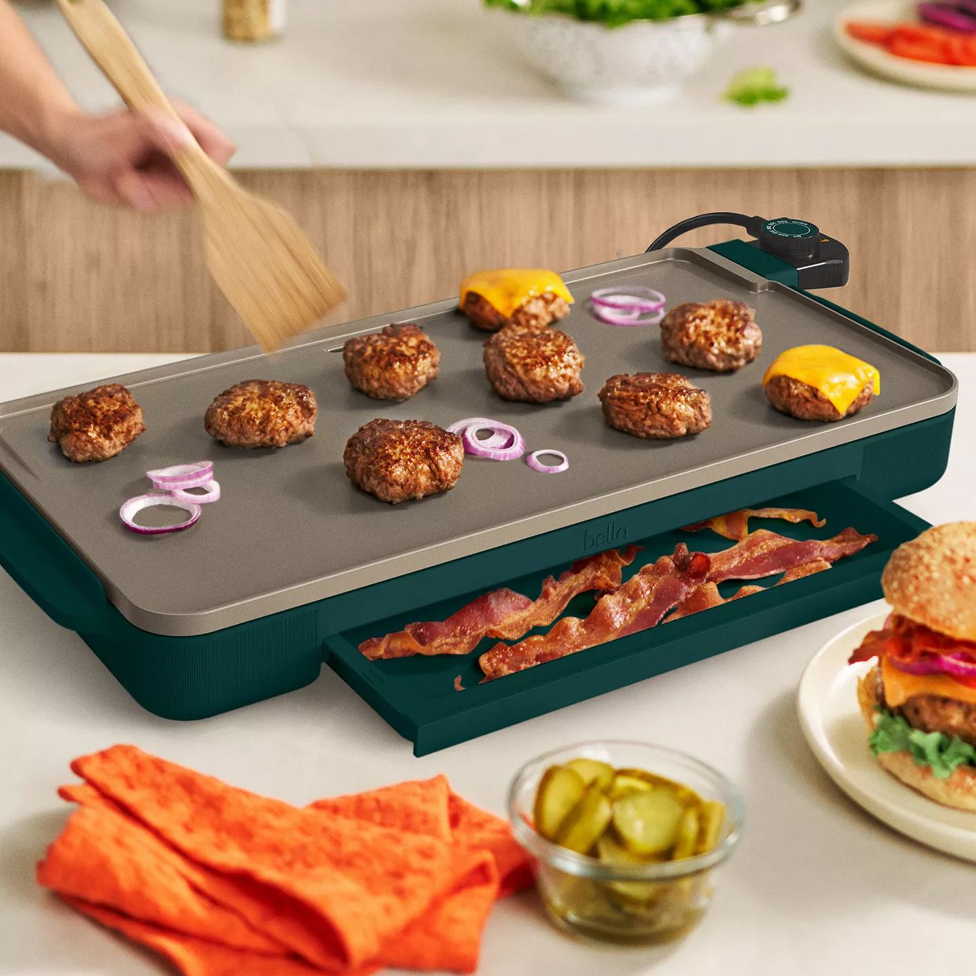 An electric griddle with sliders, onions, and bacon features a warming tray for bacon underneath, alongside toppings like pickles and a burger.