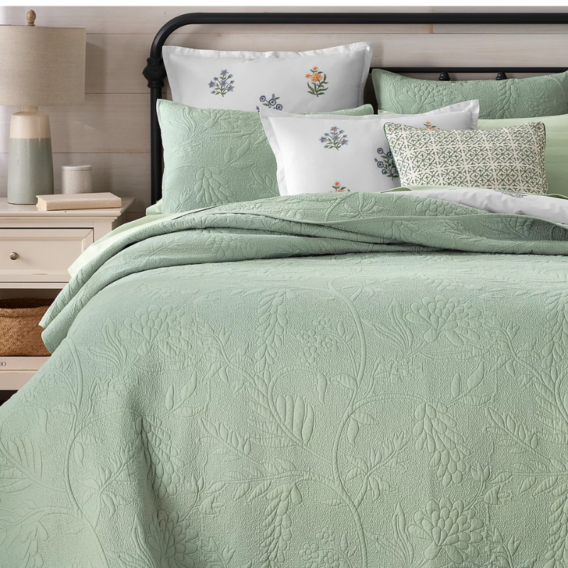 A light green quilted bedspread with floral patterns is displayed with matching pillows and a decorative cushion on a metal bed frame. The bed is accompanied by a white nightstand holding a lamp, two books, and a candle.