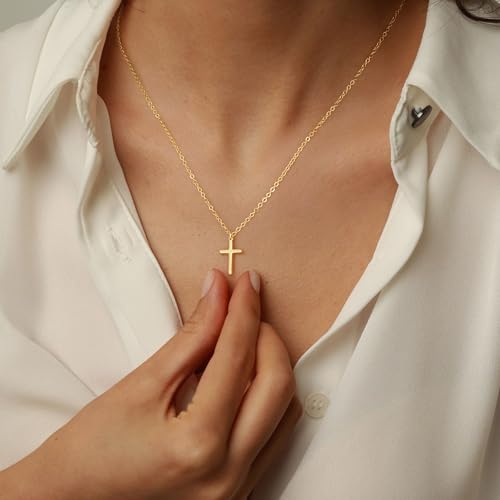A gold necklace with a cross pendant is worn by a person in a white shirt. The person is gently holding the pendant, highlighting its simple and elegant design.