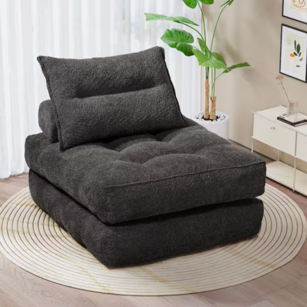 A dark gray, plush upholstered floor chair with a pillow backrest, placed on a circular striped rug in a living room setting.