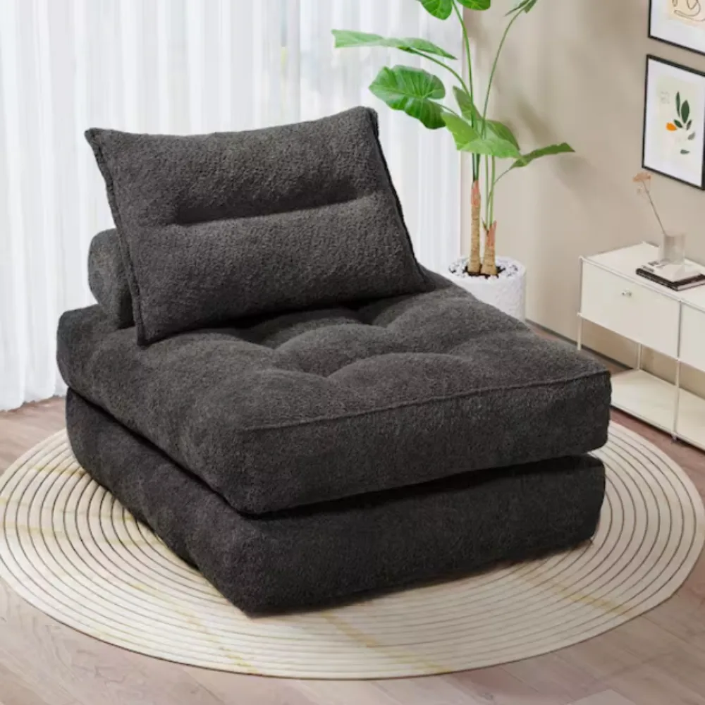 A dark gray, plush upholstered floor chair with a pillow backrest, placed on a circular striped rug in a living room setting.