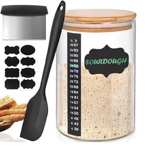 A sourdough starter jar set with a bamboo lid, temperature gauge, labels, a dough scraper, and a black spatula. Contains slices of bread.