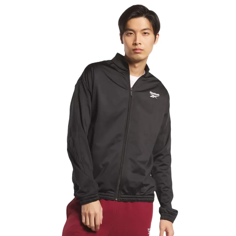 A man is wearing a black Reebok track jacket with a full zipper, ribbed cuffs, and a small white logo on the left chest.