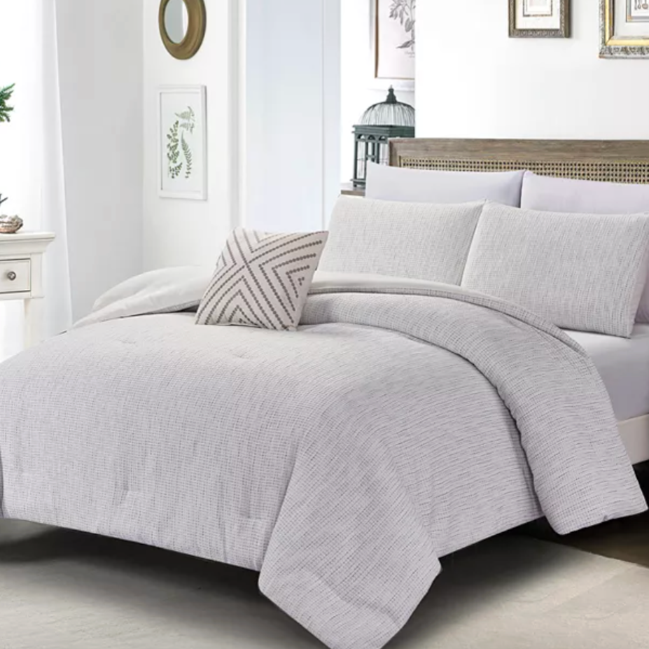 A neatly made bed features a light gray comforter with matching pillows and a decorative pillow with a geometric pattern. The setting includes a white nightstand with plants and wall decor.