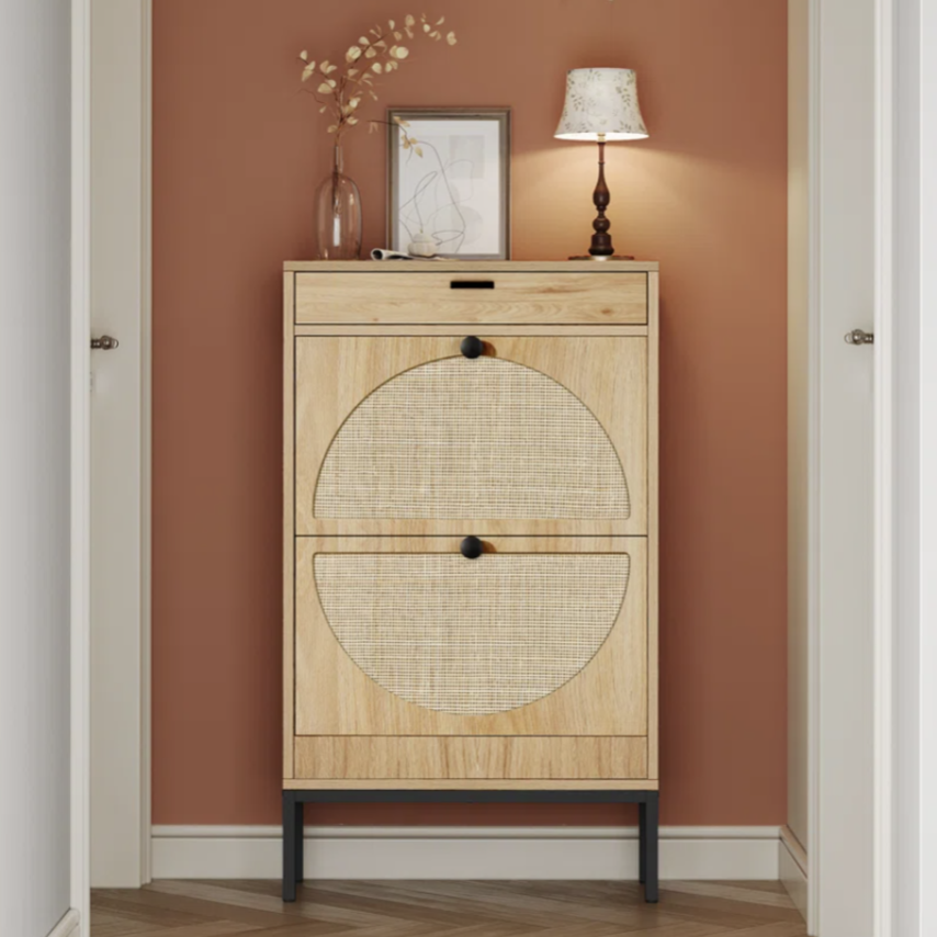A beige wooden shoe cabinet with a woven rattan design stands against a brown wall. It's topped with a lamp, decorative branch in a vase, and framed artwork, with a minimalist clock on the wall above.