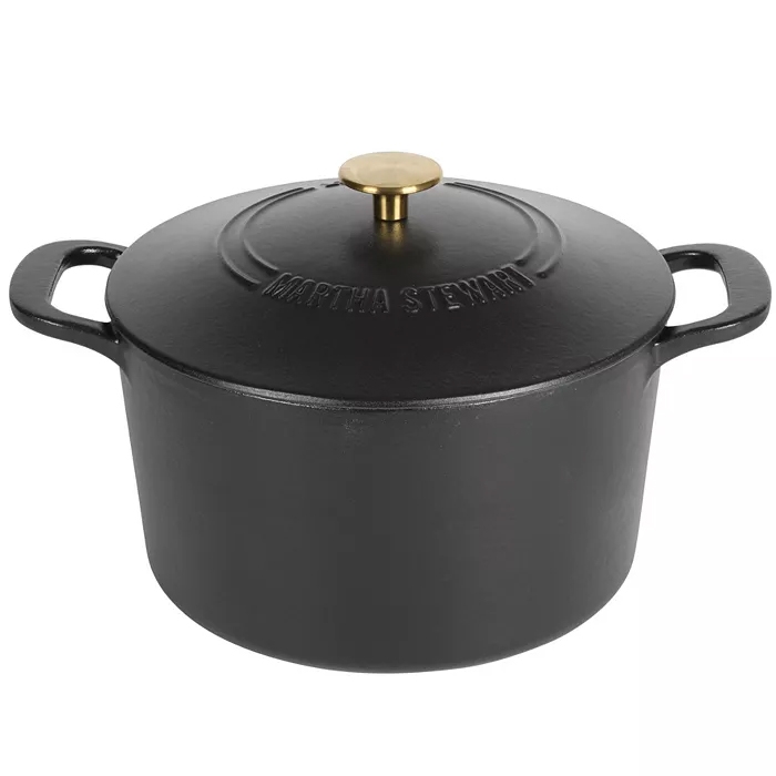 A black cast iron Dutch oven with a 7-quart capacity, featuring a lid with a brass knob and \“Martha Stewart\“ embossed on it. It has two side handles for easy handling.