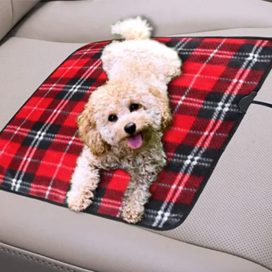 A small fluffy dog lies on a red and black plaid heating pad placed on a car seat.