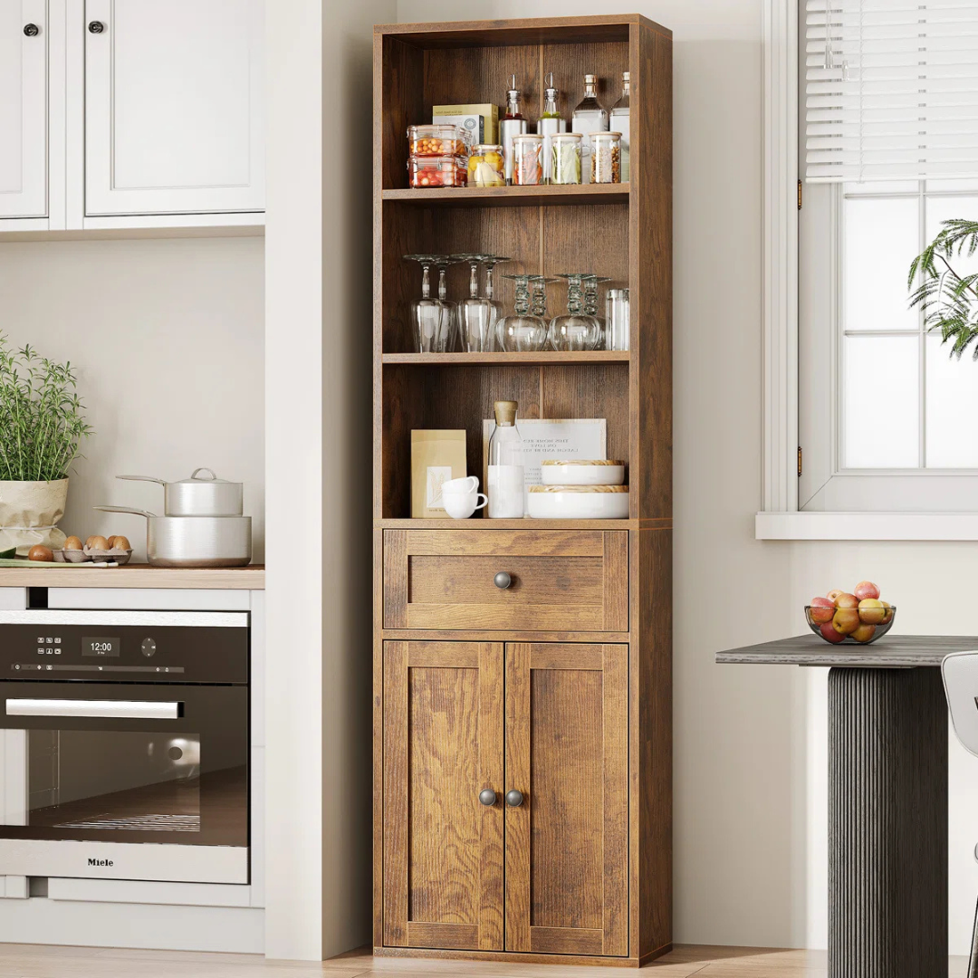 Tall wooden cabinet with upper open shelves displaying glassware and jars, a middle drawer, and lower closed storage. The setting includes a kitchen oven, a potted plant, and fruit bowl on a nearby table.
