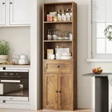 Tall wooden cabinet with upper open shelves displaying glassware and jars, a middle drawer, and lower closed storage. The setting includes a kitchen oven, a potted plant, and fruit bowl on a nearby table.