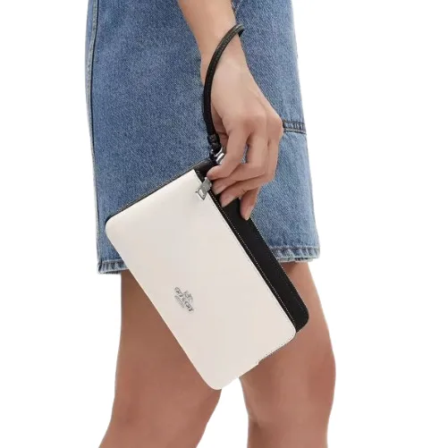 A person is holding a white zippered wristlet with a black accent and a logo, against a background of a denim skirt.