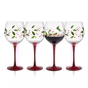 Set of four wine glasses with red stems, adorned with holly and berry designs. One glass is filled with red wine.