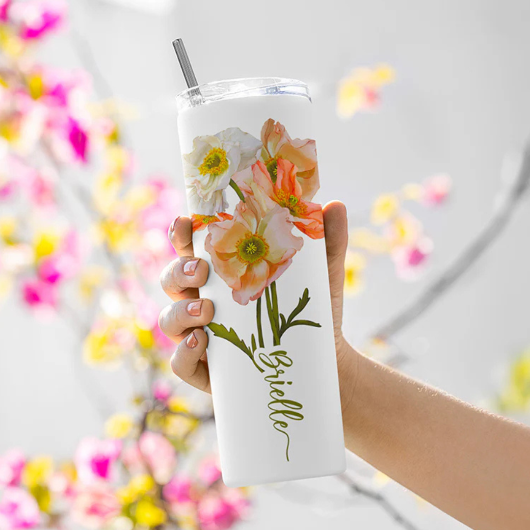 A hand holds a slim tumbler with floral designs and a metal straw. The tumbler features orange and white flowers with the name \“Brielle\“ written in cursive. The background shows blurred pink and yellow flowers.