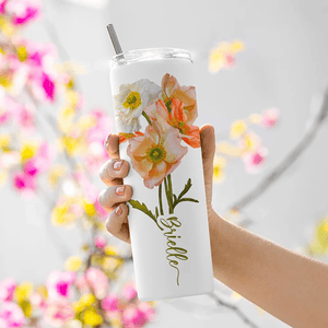 A hand holds a slim tumbler with floral designs and a metal straw. The tumbler features orange and white flowers with the name \“Brielle\“ written in cursive. The background shows blurred pink and yellow flowers.
