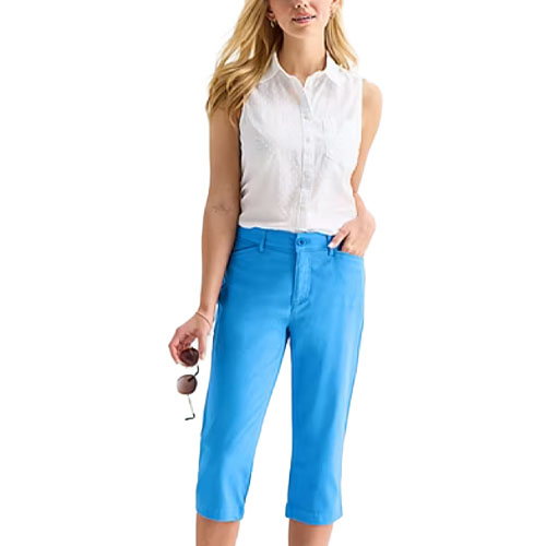 A woman is wearing a white sleeveless button-up blouse and bright blue knee-length pants, holding sunglasses in her right hand.
