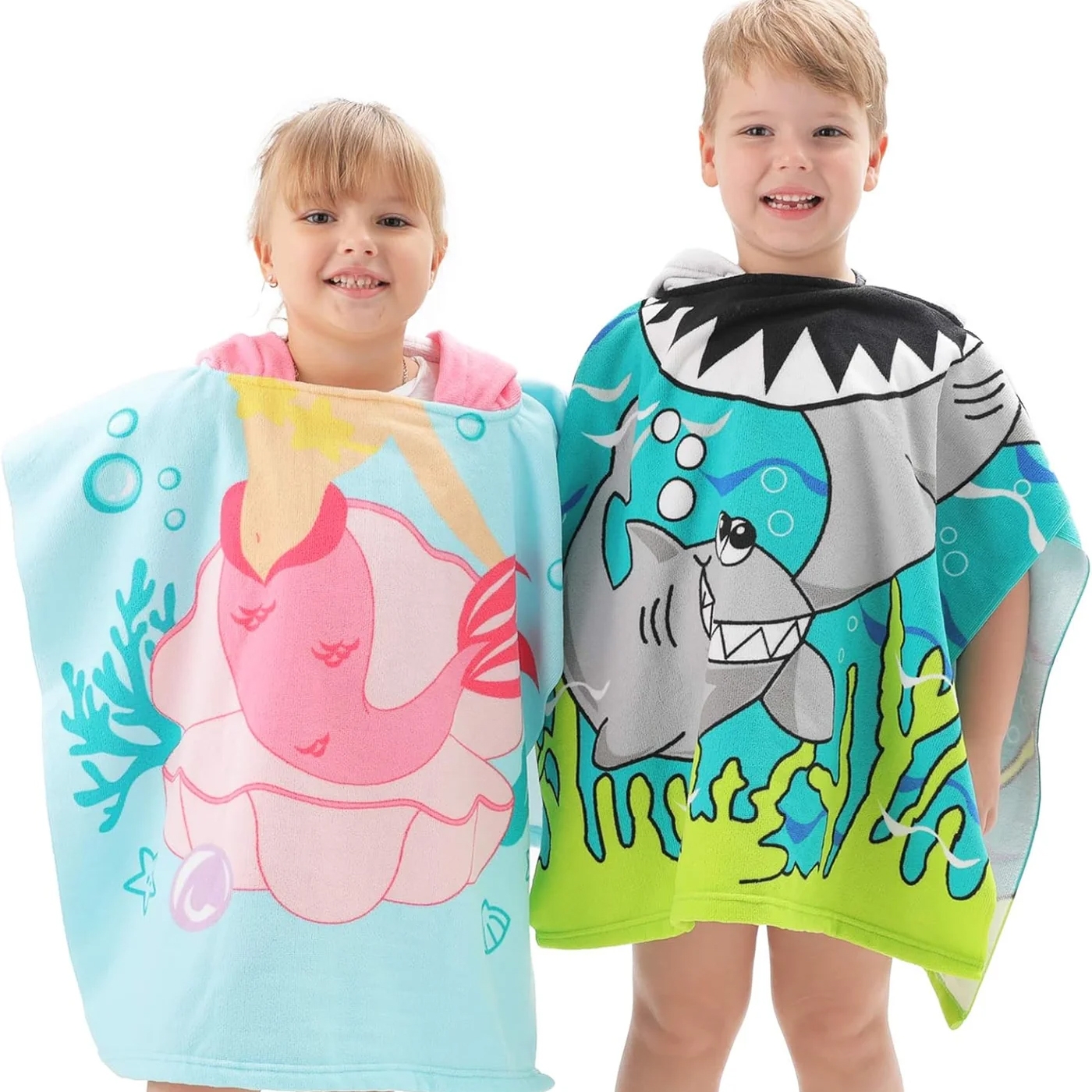 Two children are wrapped in hooded bath towels, one with a pink mermaid design and the other with a green shark design.