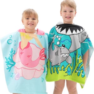 Two children are wrapped in hooded bath towels, one with a pink mermaid design and the other with a green shark design.