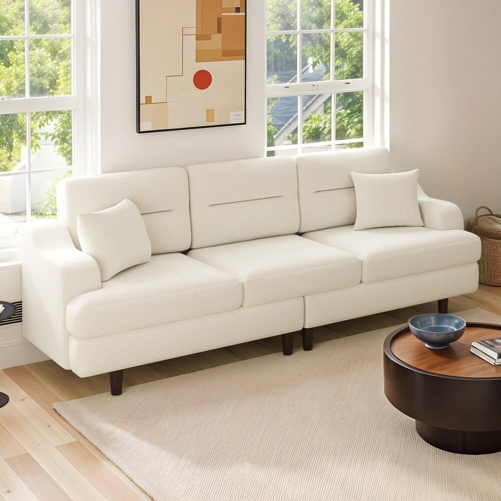 A modern white sofa with cushions is placed in a bright living room, accompanied by a round wooden coffee table and a minimalist wall art piece.