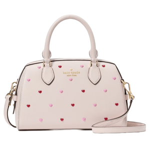 Cream-colored duffle bag with pink and red heart studs, featuring dual handles, gold hardware, and a detachable shoulder strap. The brand name \“Kate Spade New York\“ is embossed in gold on the front.