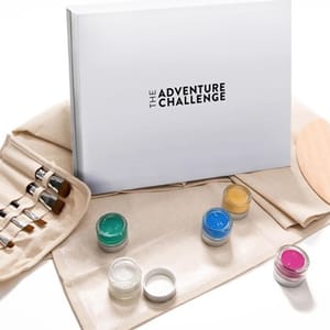 An \“Adventure Challenge\“ book is displayed with paintbrushes, jars of colorful paint, and a palette on a cloth surface.