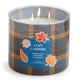 A triple wick candle labeled \“Cozy Campfire\“ with scents of autumn woods, tonka bean, and eucalyptus, featuring soy wax and essential oils. The packaging has a plaid design with autumn leaf illustrations.