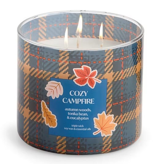A triple wick candle labeled \“Cozy Campfire\“ with scents of autumn woods, tonka bean, and eucalyptus, featuring soy wax and essential oils. The packaging has a plaid design with autumn leaf illustrations.