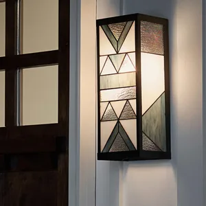 A rectangular stained glass wall sconce with geometric patterns, featuring stained glass panels in shades of blue and white with a black metal frame, is mounted on a wall beside a wooden door.