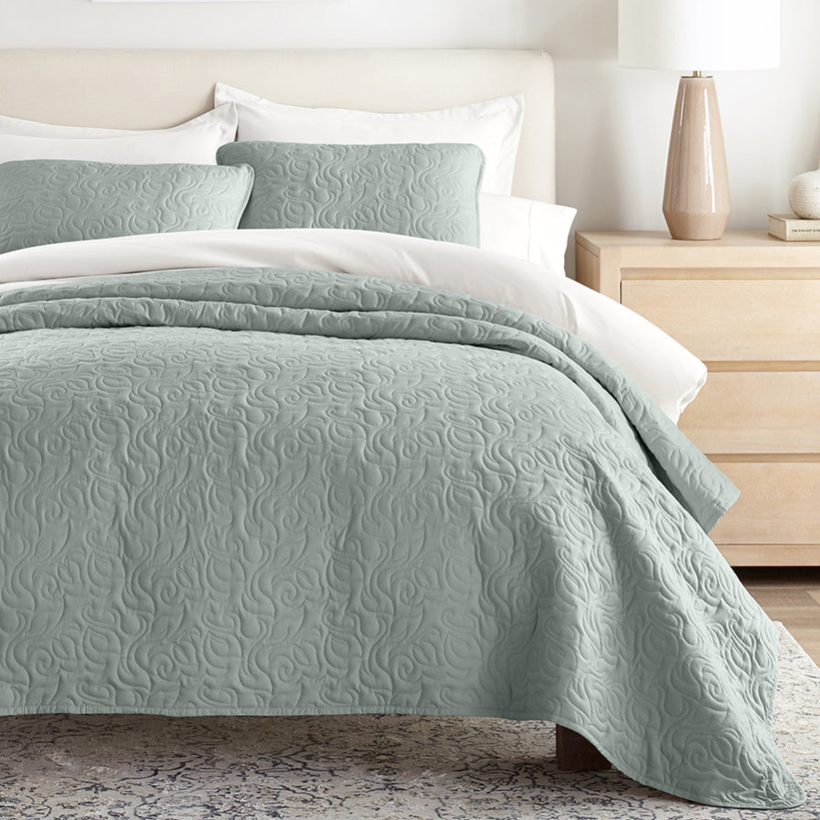 A bedroom with a pastel green quilt featuring a swirling pattern, two matching pillows, and a light wood dresser with a ceramic lamp.
