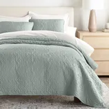 A bedroom with a pastel green quilt featuring a swirling pattern, two matching pillows, and a light wood dresser with a ceramic lamp.